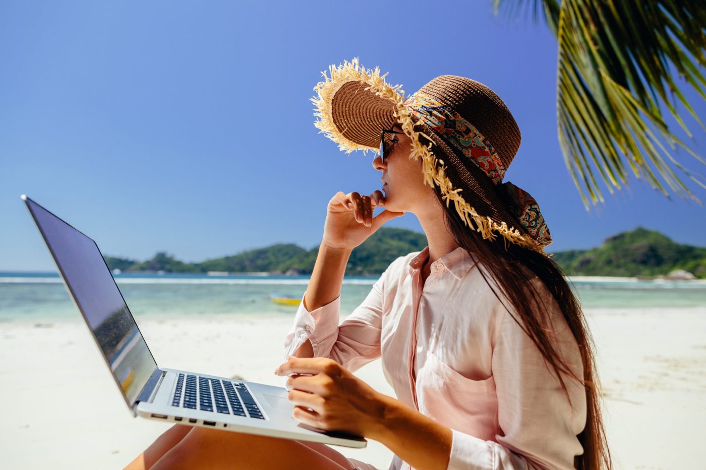 A traveler in a sun hat working on a laptop on a tropical beach